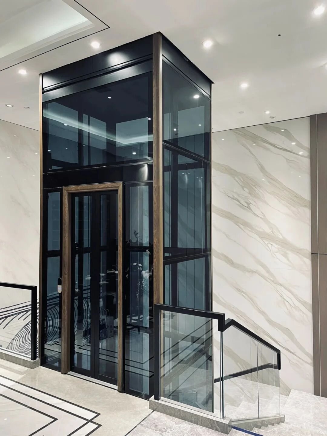 Levitex Max glass lift with a dark frame and bronze door trim beside a marble wall.